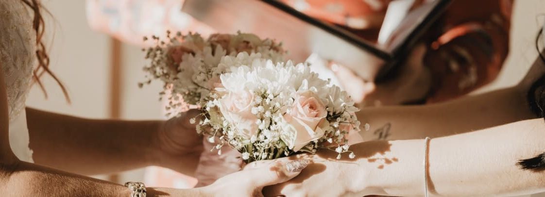 Close-up of two people holding a bouquet of light pink and white flowers during a wedding ceremony.