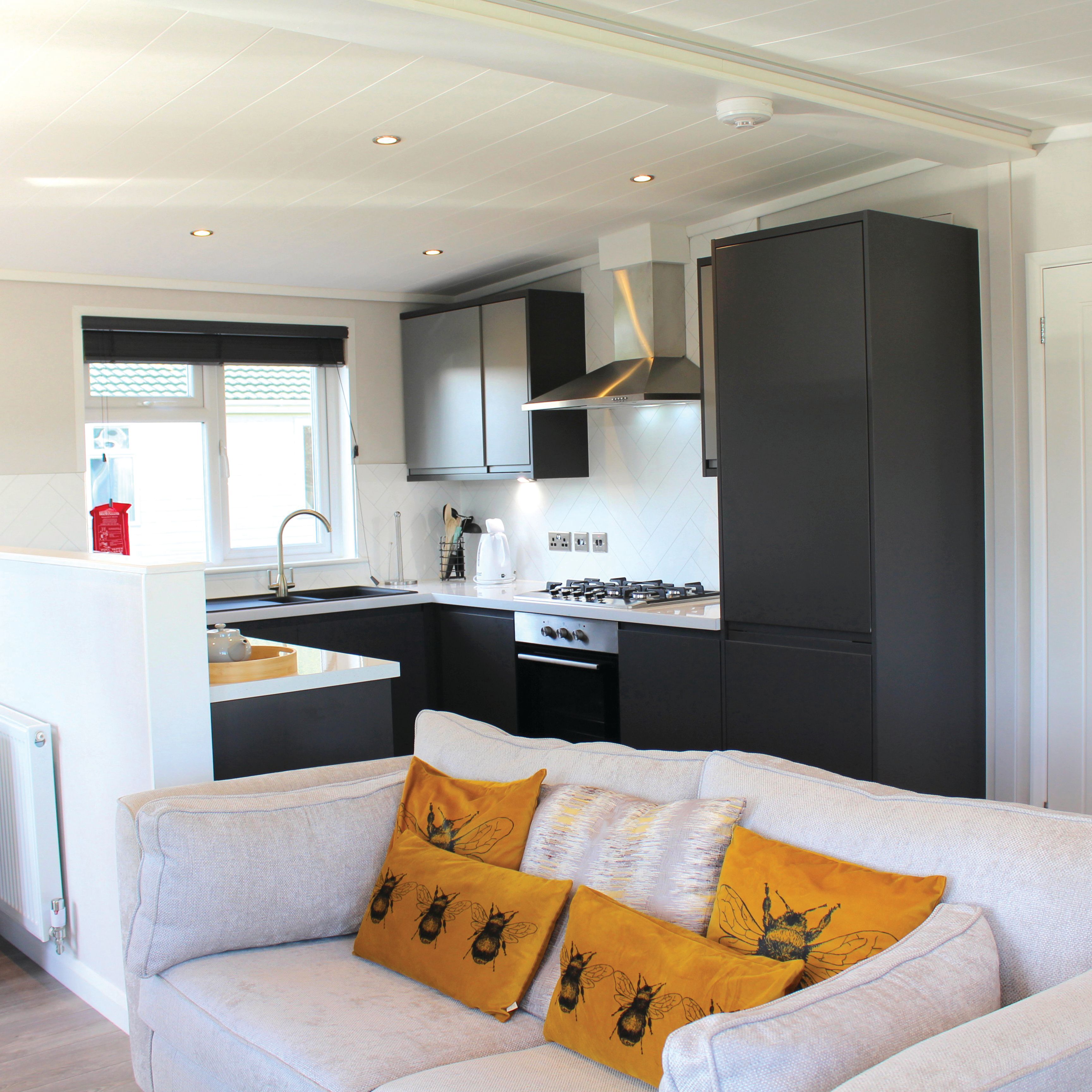 Modern open-plan living area with grey sofas, orange bee-themed cushions, and a stylish kitchen with black cabinets.