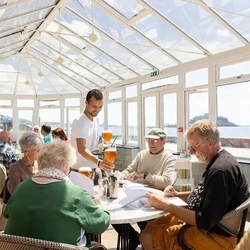 Waiter serving drinks to a group of elderly people at a sunlit seaside restaurant
