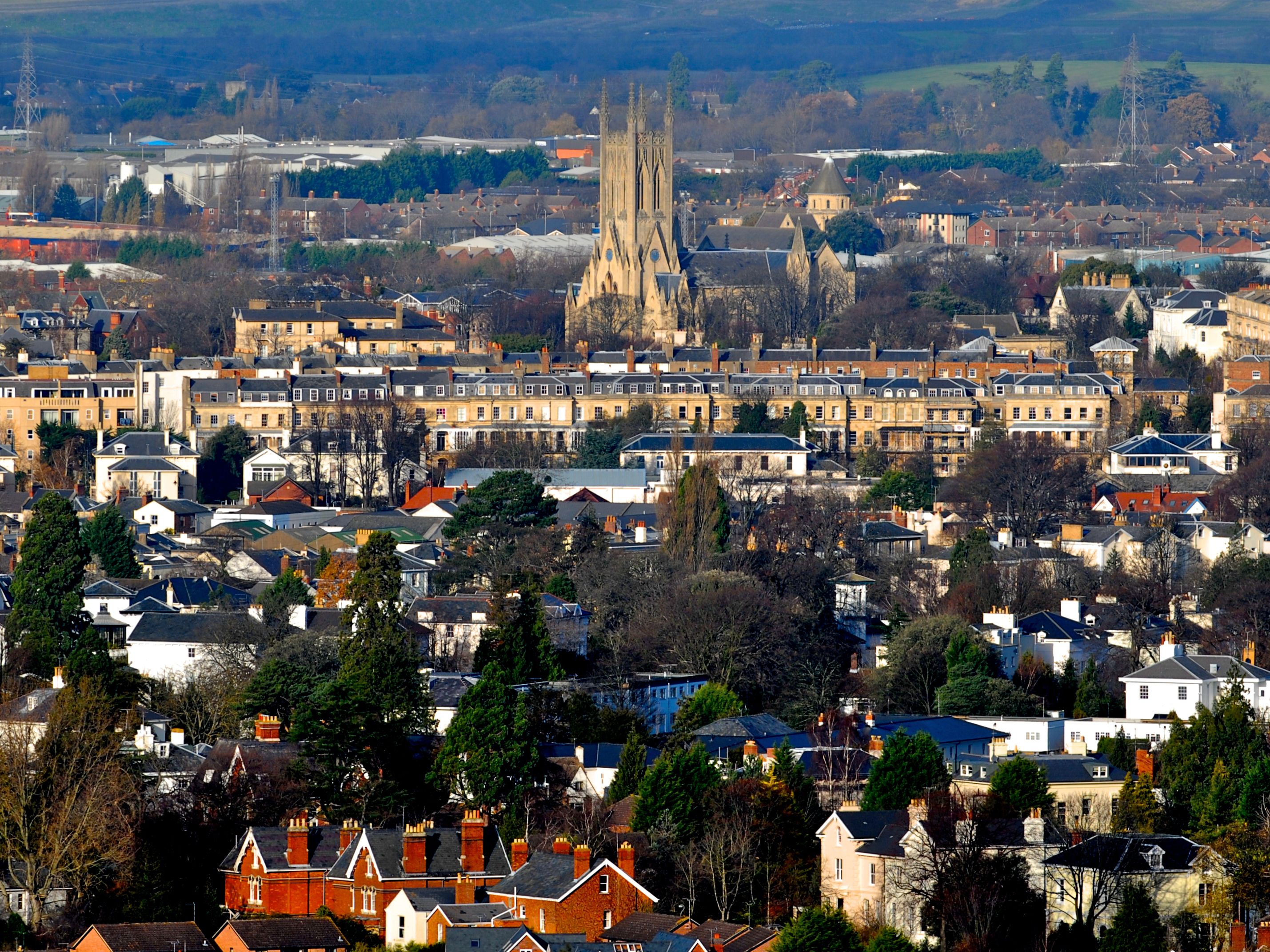 A cityscape featuring a mix of residential and historic buildings, with a Gothic-style cathedral dominating the skyline, surrounded by trees and rolling hills in the background.