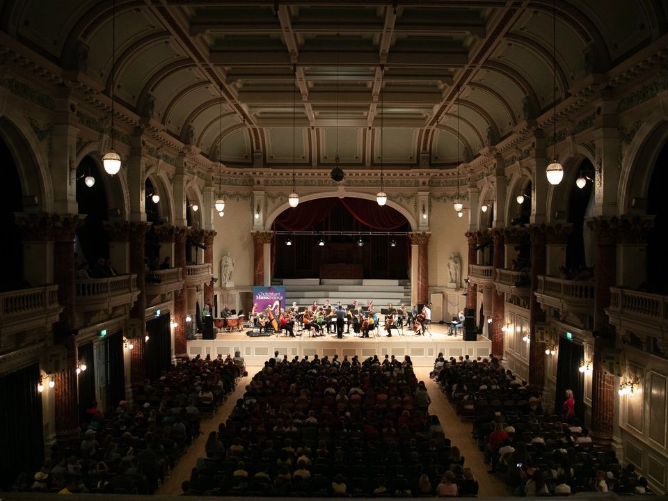 A classical orchestra performs on stage in a grand concert hall with ornate architecture, high ceilings, and decorative lighting, as an audience watches attentively.