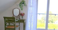 Vintage-style vanity table and chair by a large window with floral decor and a hanging plant.
