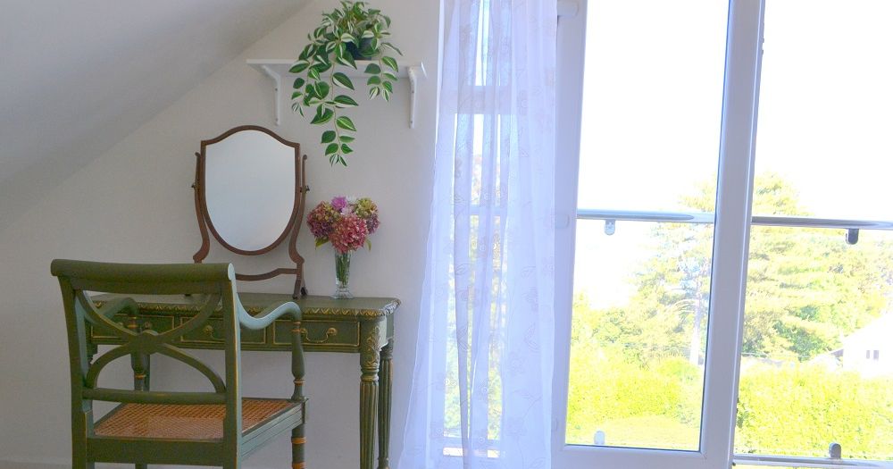 Vintage-style vanity table and chair by a large window with floral decor and a hanging plant.