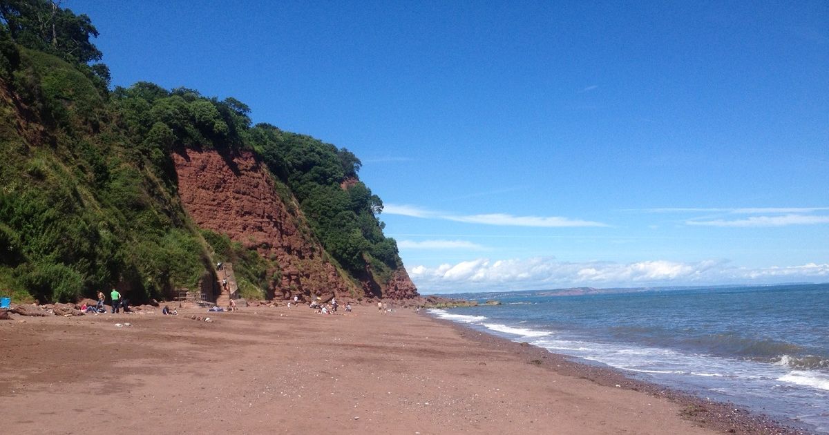 Ness Cove beach in Shaldon with red cliffs, sandy shore, and calm blue sea under a clear sky
