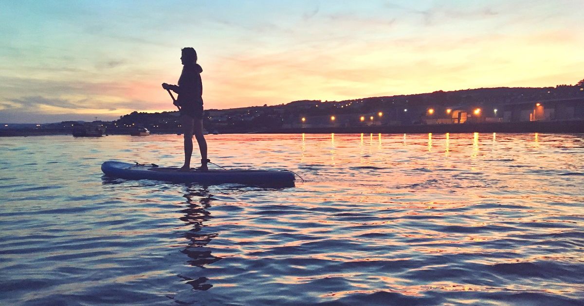 Person stand-up paddleboarding on a calm body of water at sunset.