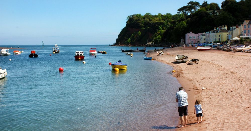 Shaldon beach with boats on the water, colorful houses, and a man walking with a child along the sandy shore.