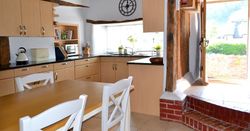 Rustic kitchen with wooden beams, light wood cabinets, a dining table with white chairs, and an open door leading outside.