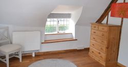 A cozy attic room with wooden beams, a small window seat, a wooden chest of drawers, and a cushioned chair.