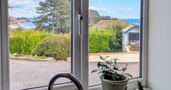 View through a kitchen window showing a garden, trees, a distant coastline with cliffs, and blue sea under a partly cloudy sky.