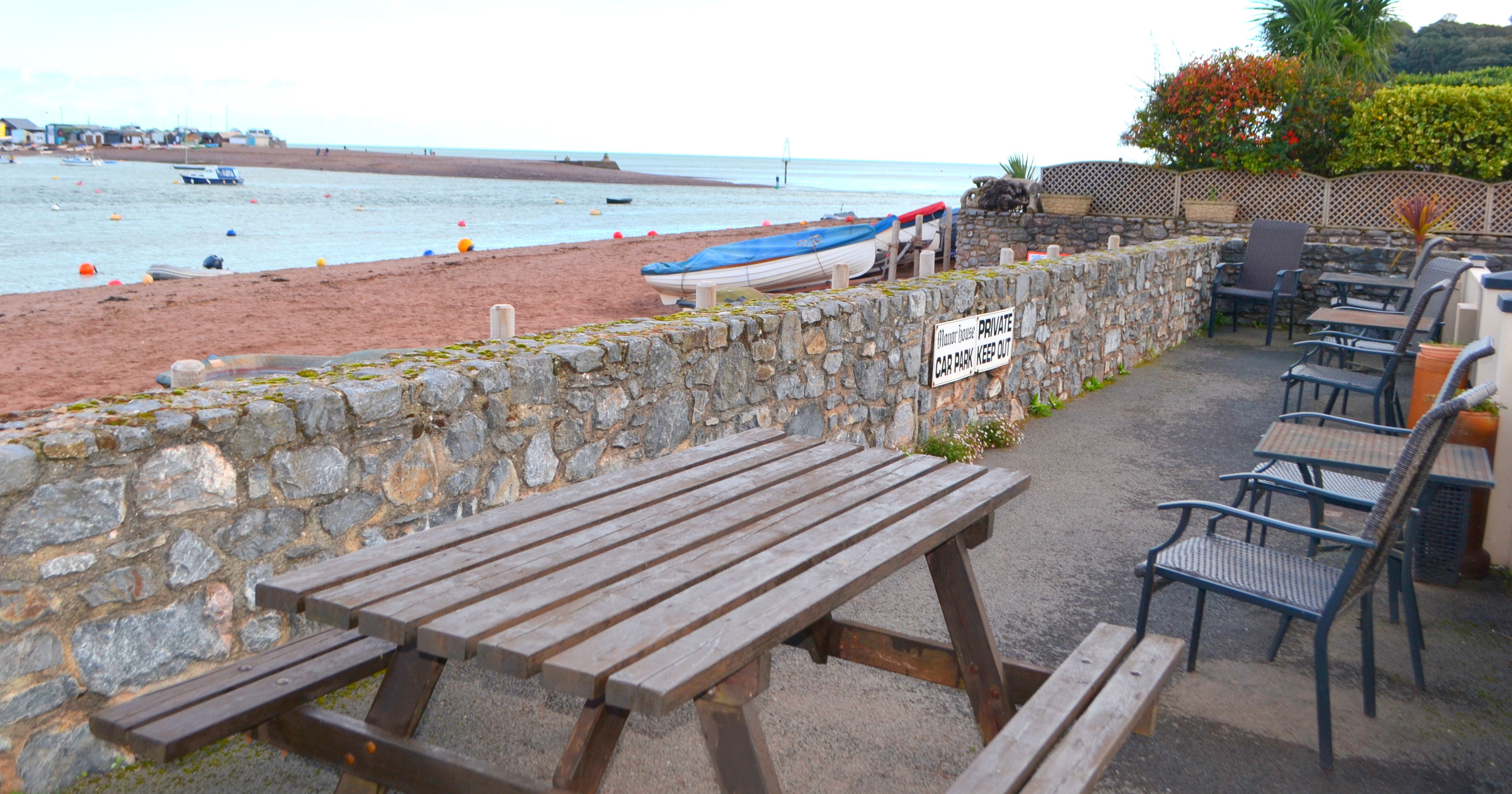 Seaside terrace with picnic table, chairs, stone wall, and view of sandy beach with boats and buoys.