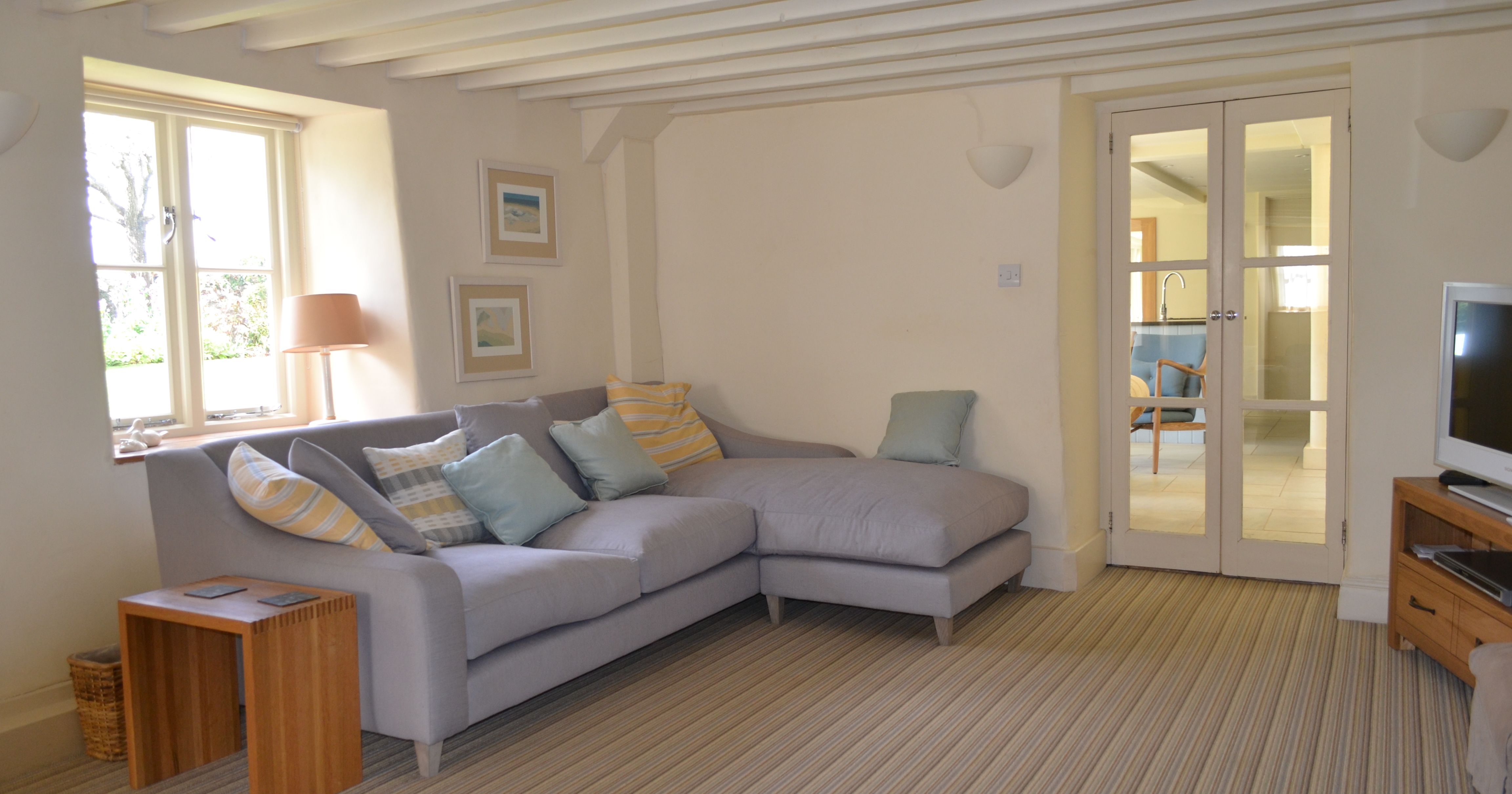 Cozy living room with a grey sectional sofa, pastel cushions, striped carpet, and French doors leading to a kitchen area.