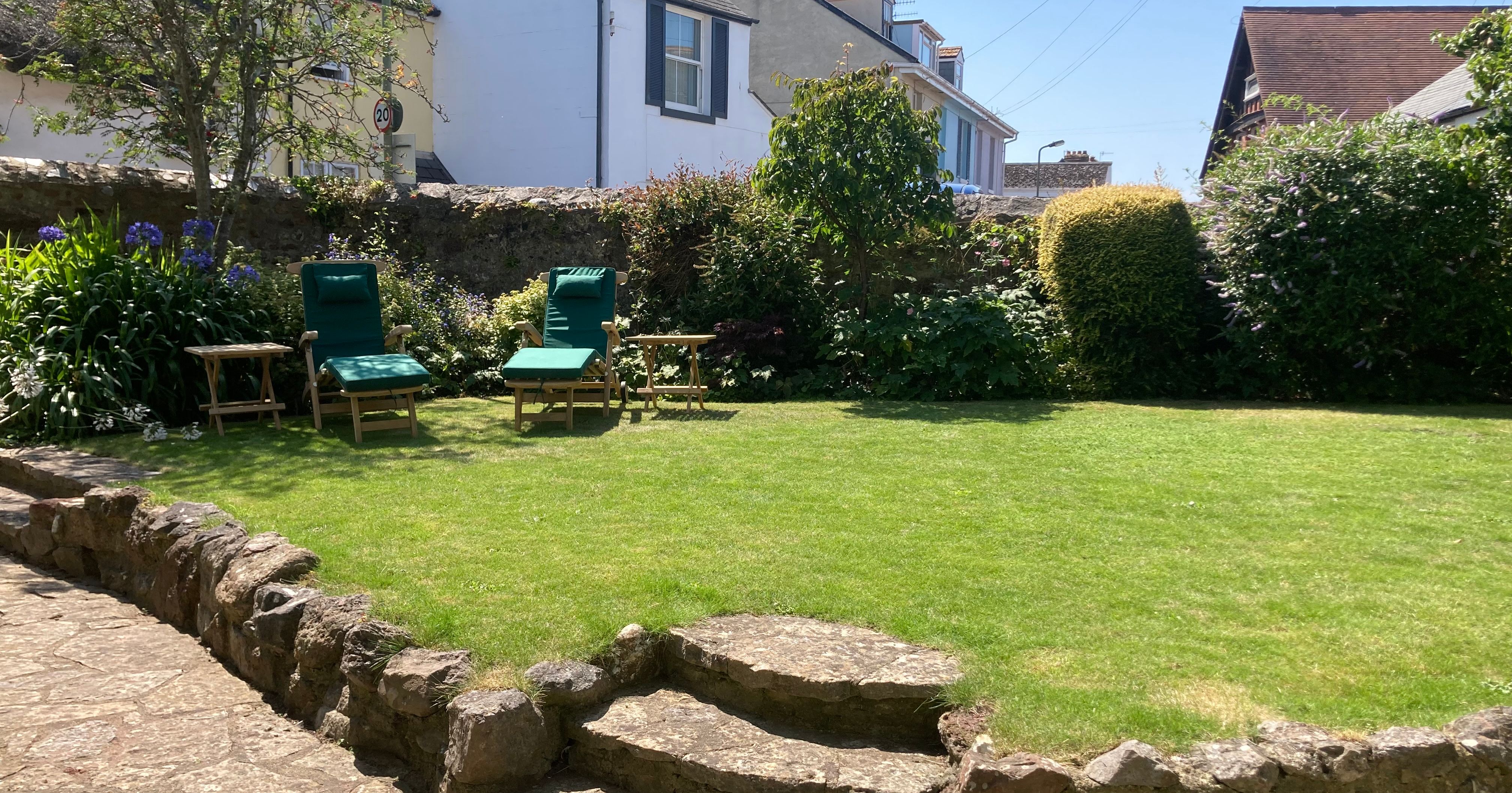 Sunny garden with two green chairs and stone steps, surrounded by plants and houses