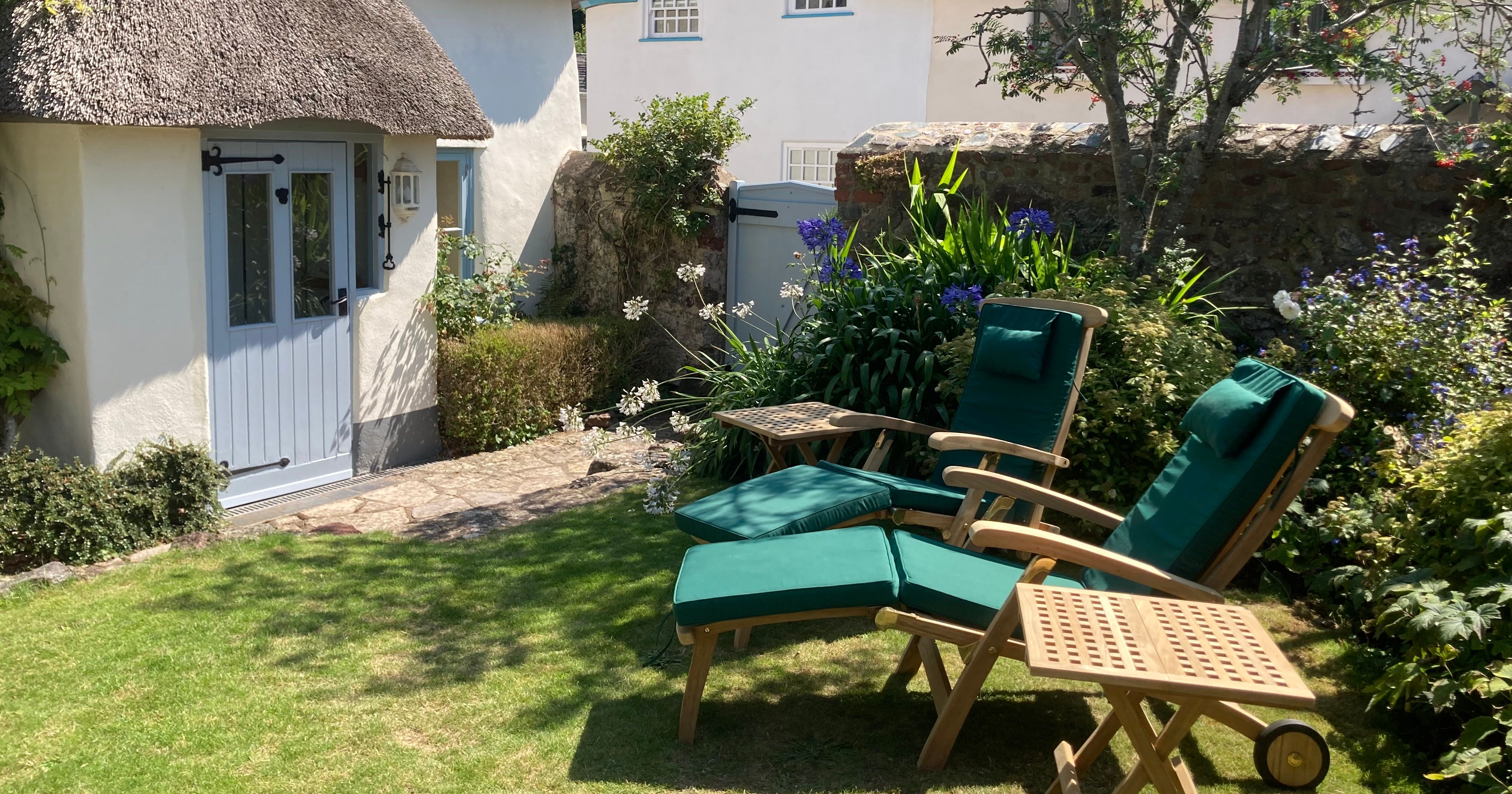Thatched cottage with a garden, two lounge chairs, and summer flowers.