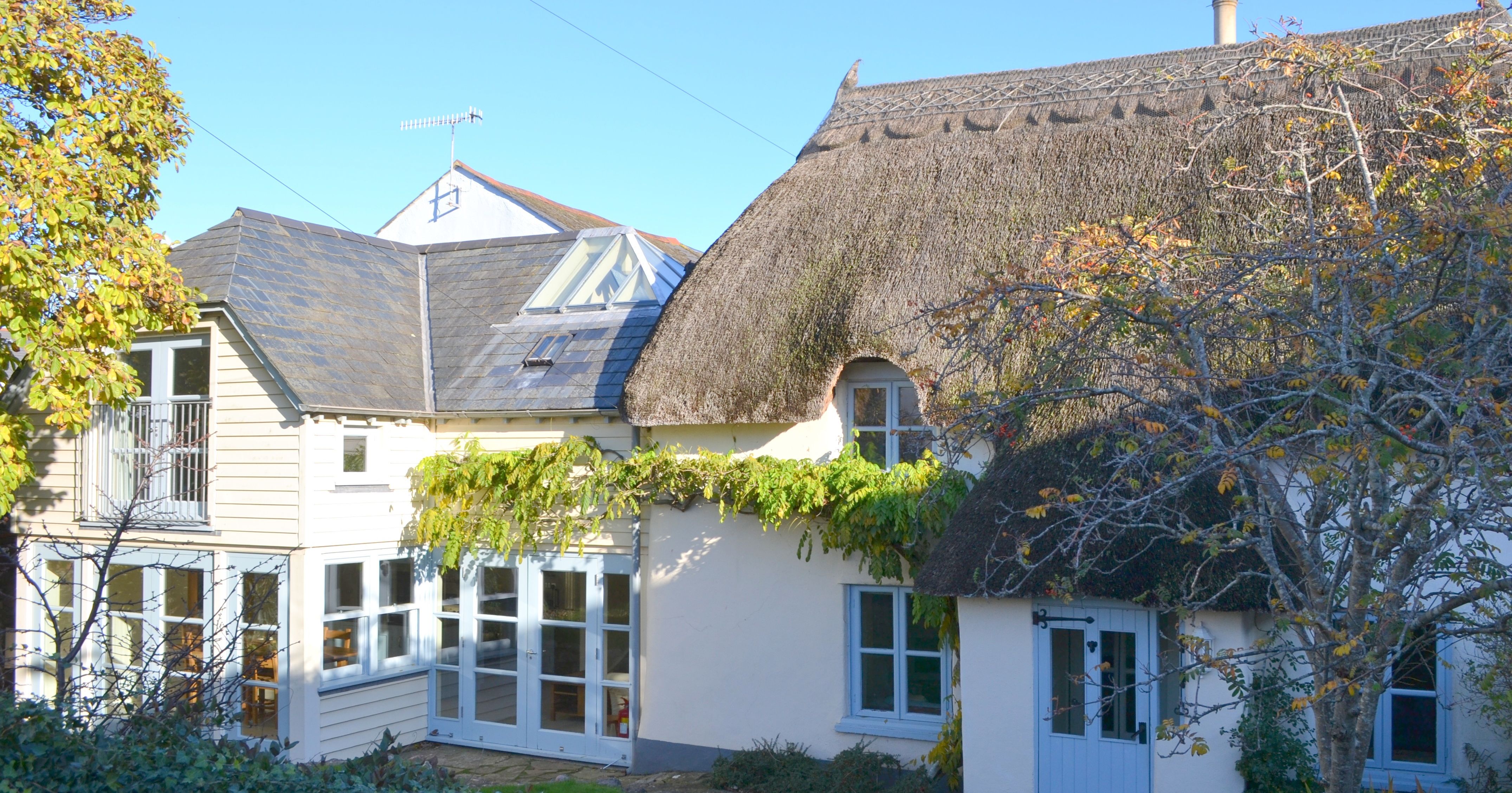 Traditional English cottage with thatched roof and white exterior surrounded by greenery.