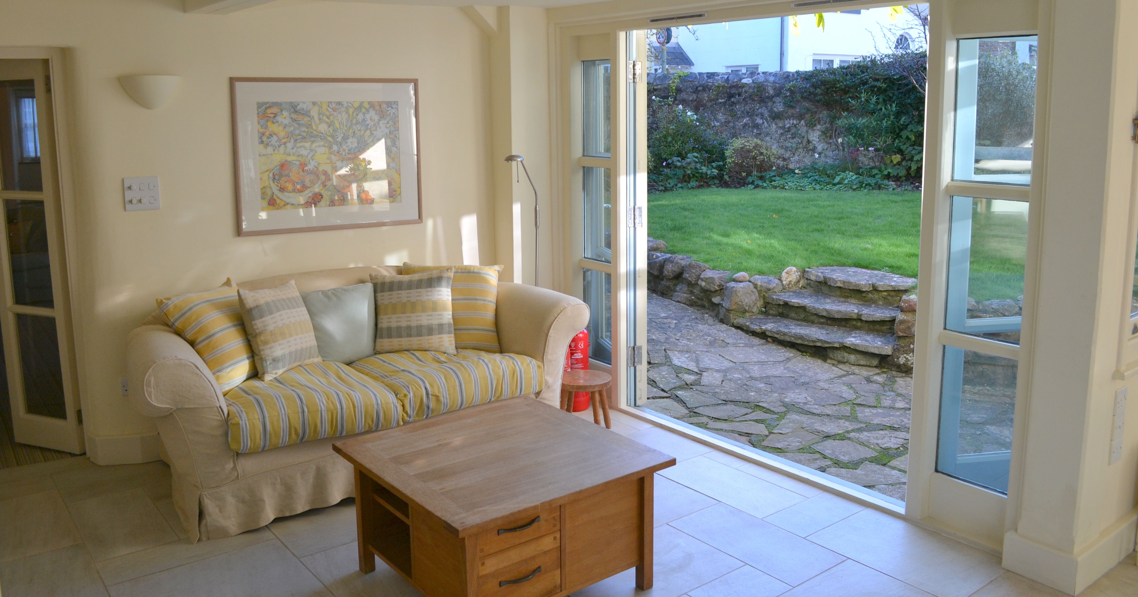 A cozy living room with a beige sofa, striped cushions, wooden coffee table, and open glass doors leading to a green garden with stone steps.