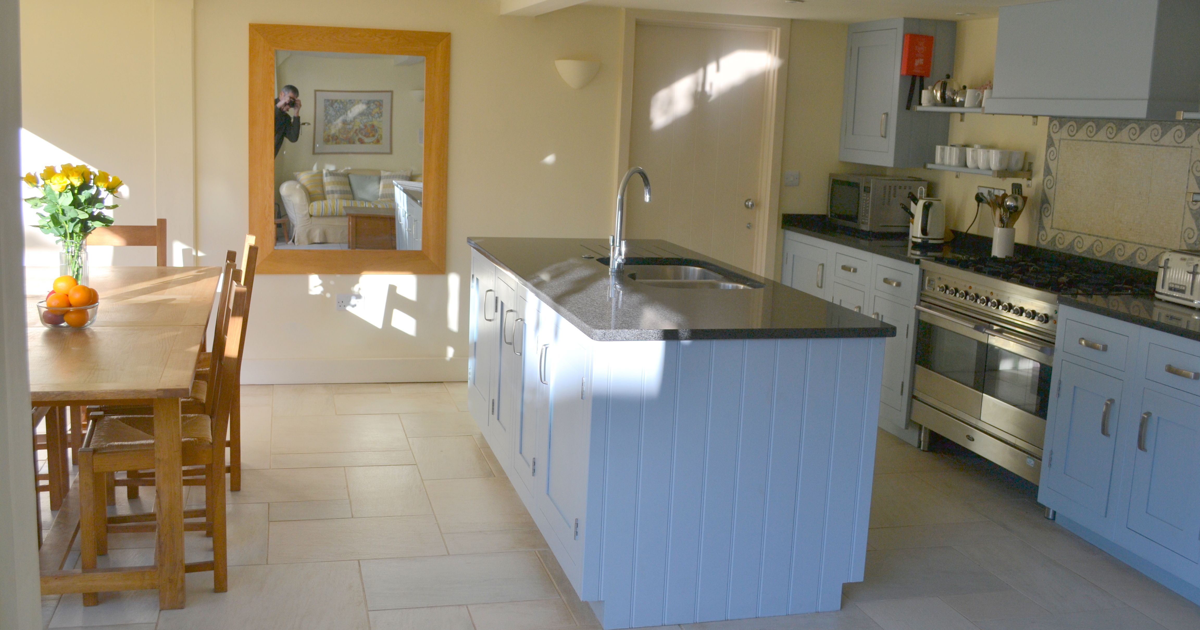 A modern kitchen and dining area with a wooden table, blue cabinets, and a large wall mirror reflecting a sitting room.