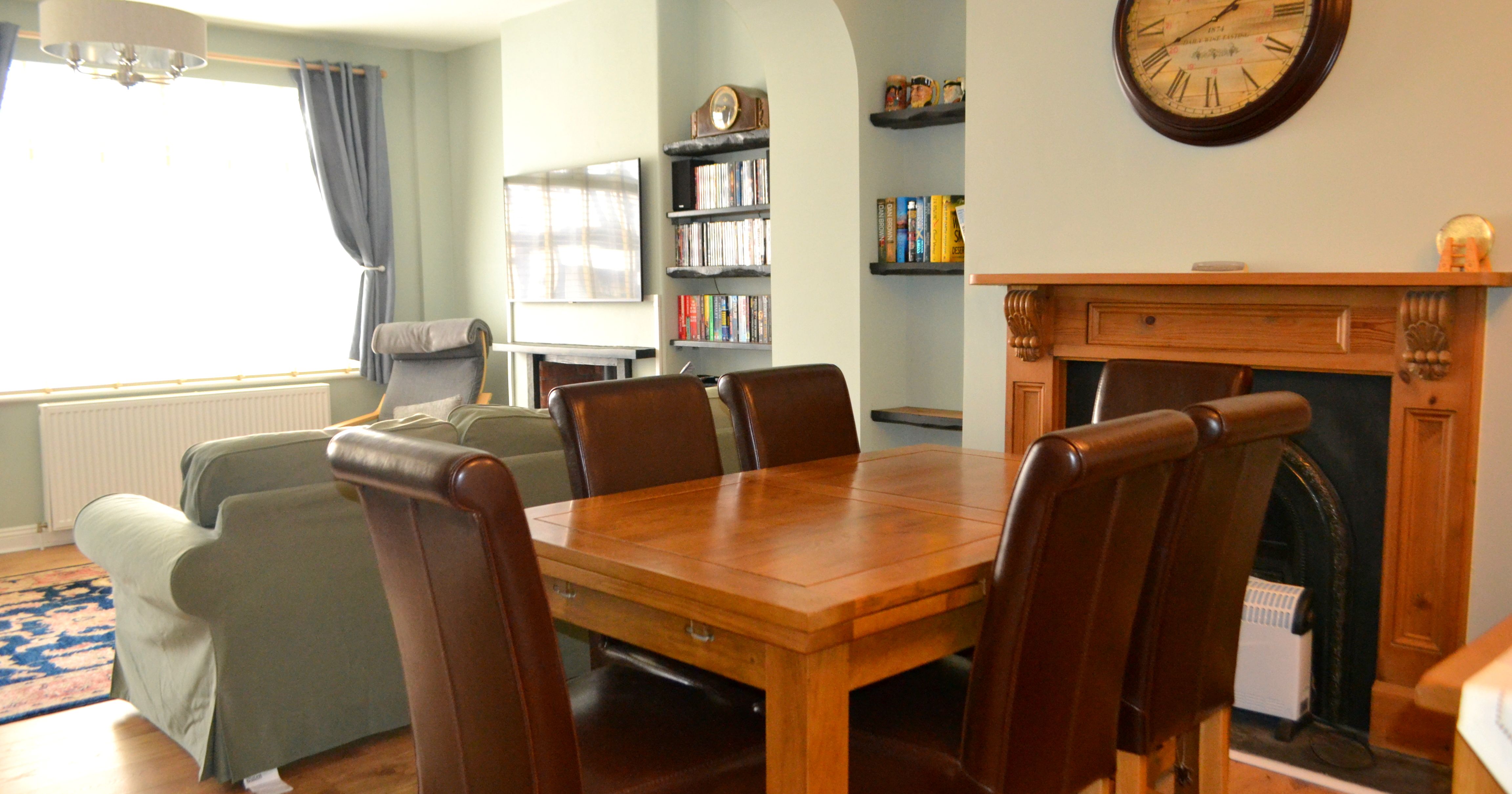 A cozy living and dining room featuring a wooden dining table with brown chairs, a fireplace with a clock above, shelves with books, sofas, and a window with curtains.
