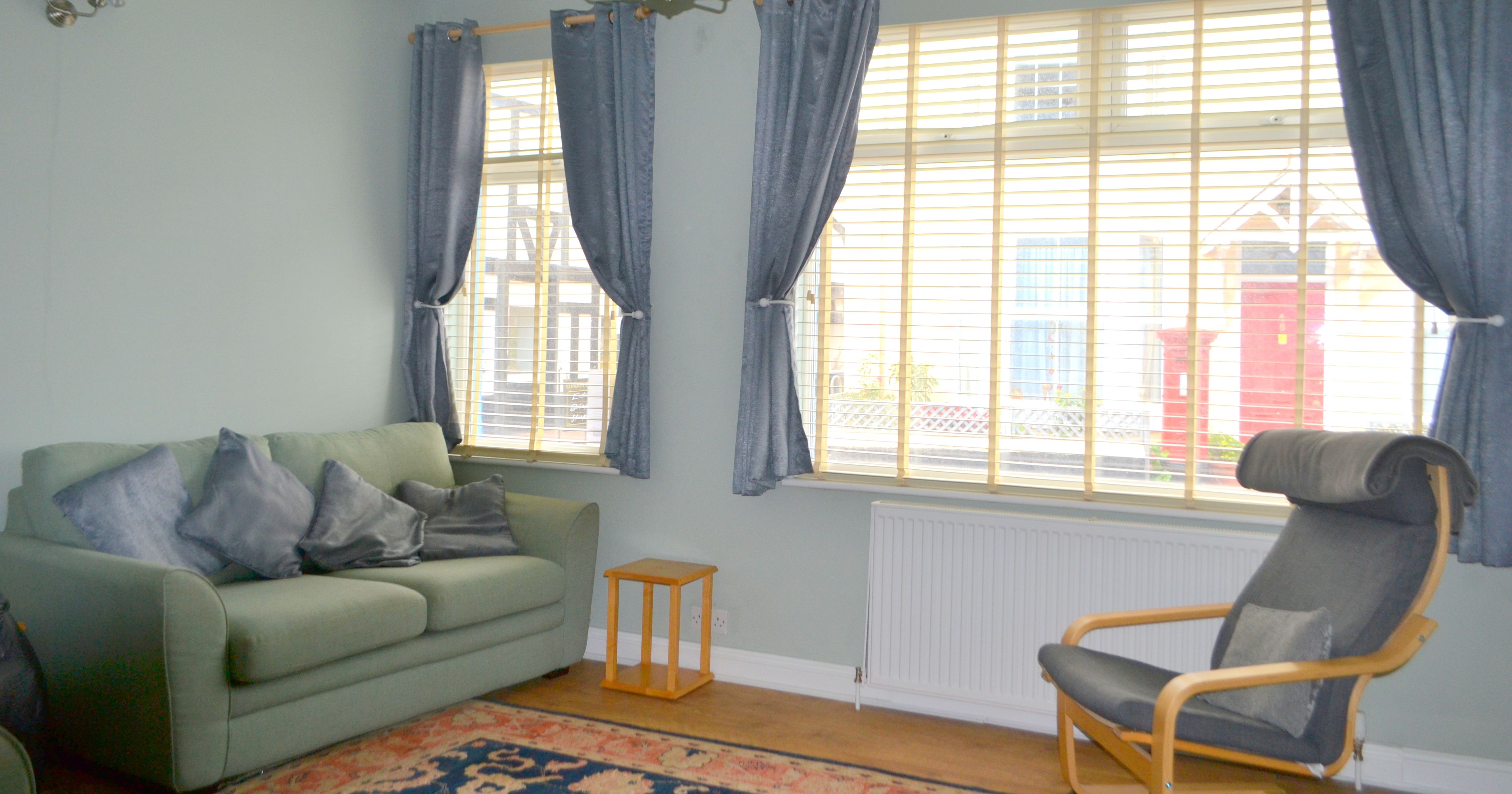 Bright living room with a green sofa, blue curtains, large window blinds, and a black armchair.