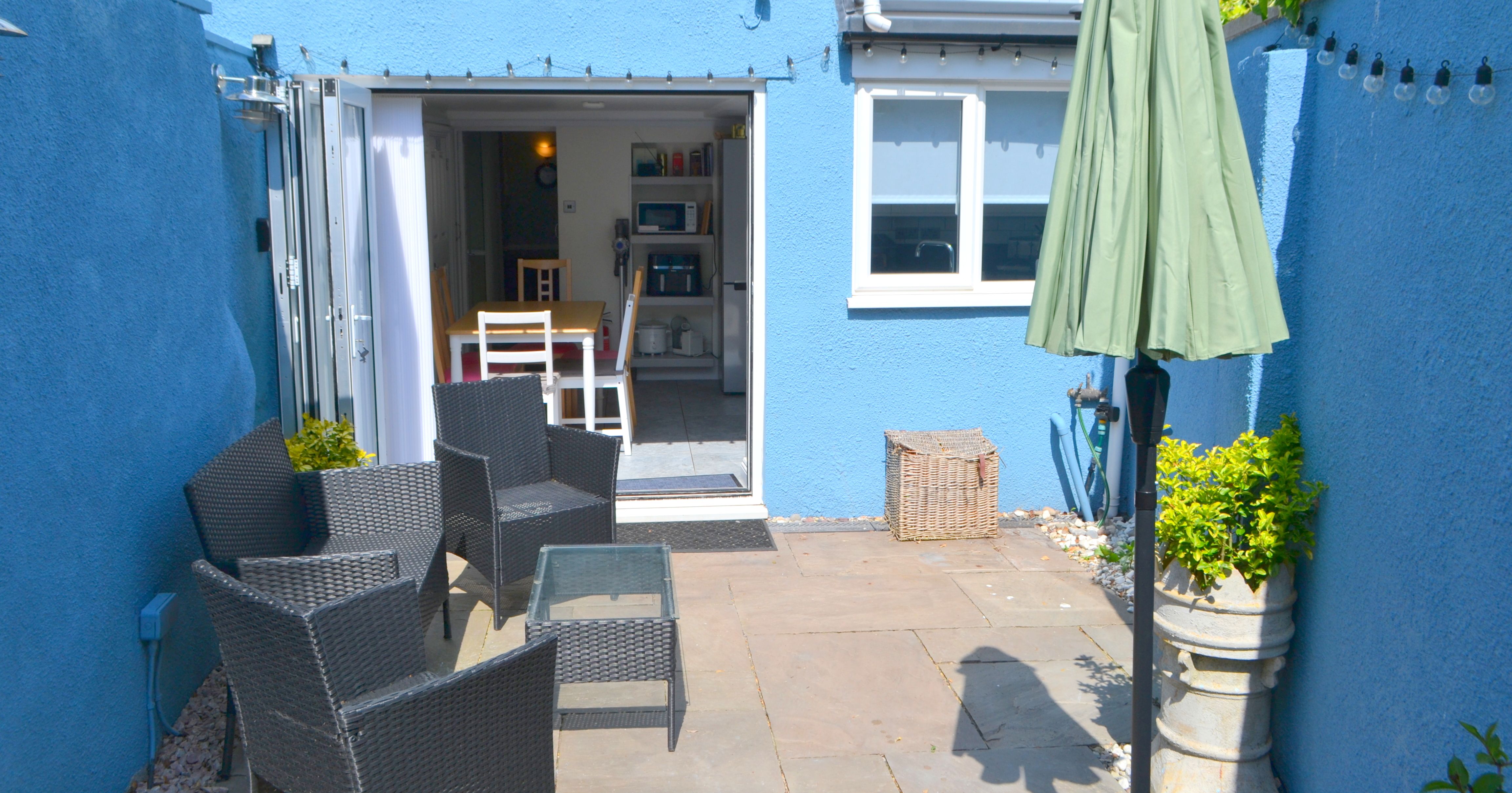 Small blue-walled backyard patio with black rattan furniture and a closed green patio umbrella.
