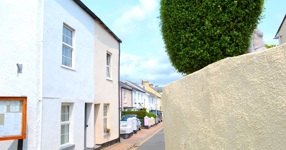 A small street lined with pastel-painted houses, a textured wall, and a trimmed tree.