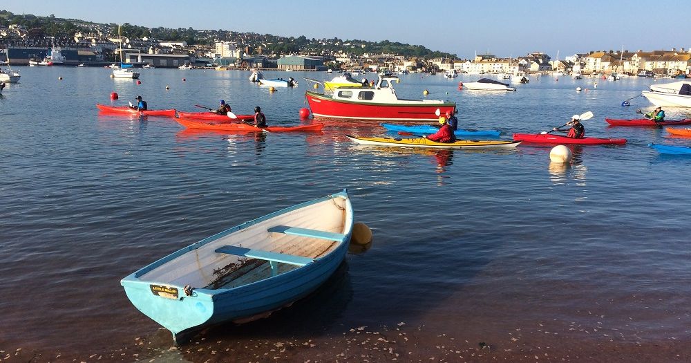 A group of people paddling canoes and kayaks on a calm harbor with various boats and a town in the background.