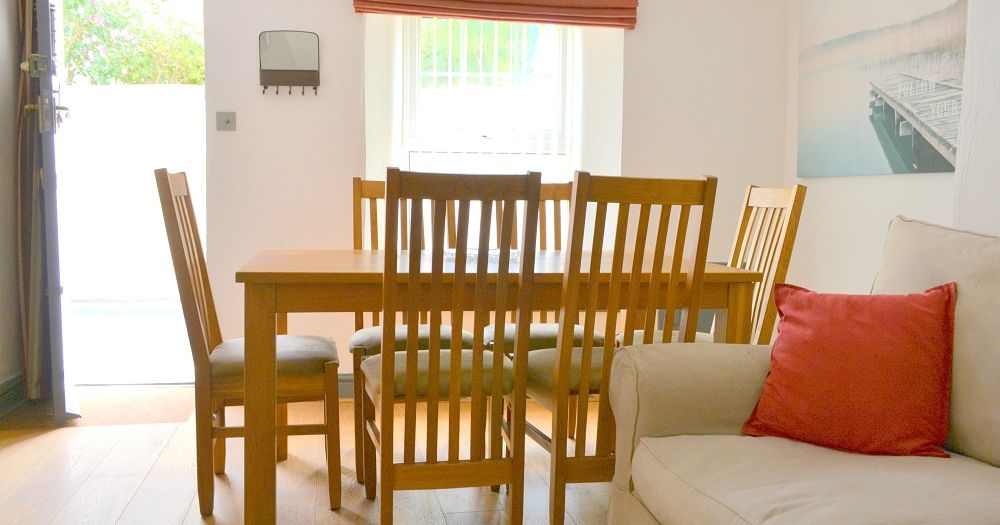 Bright dining area with wooden table and chairs, sofa with red cushion, window with red shade, and wall art.