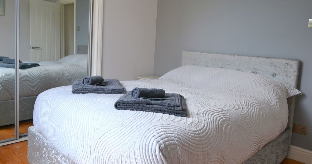 Modern double bedroom with neatly made bed, grey headboard, and folded towels on top.