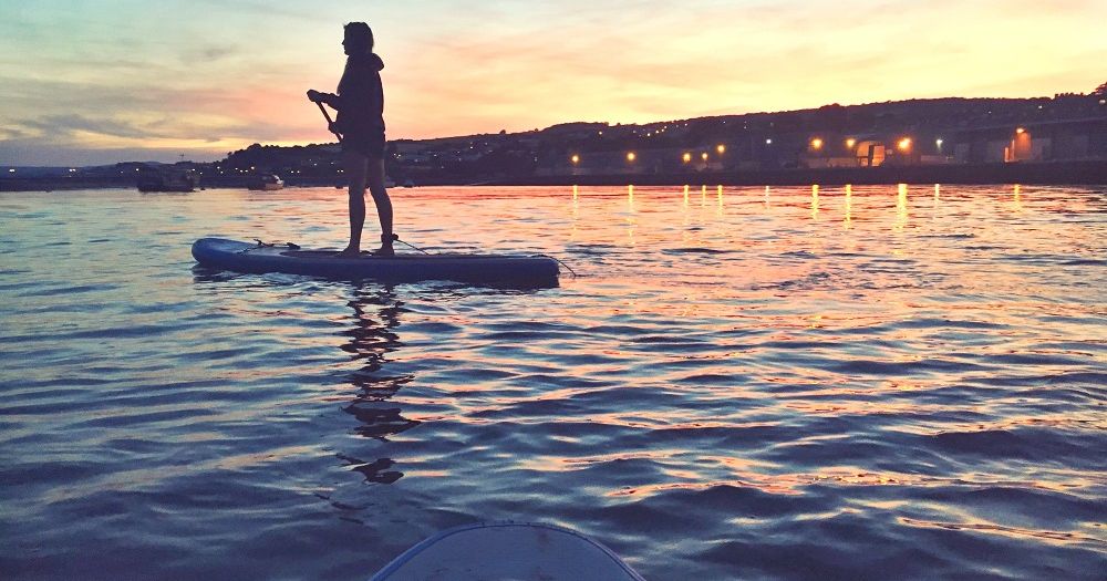 Person paddleboarding on calm water at sunset with city lights in the background