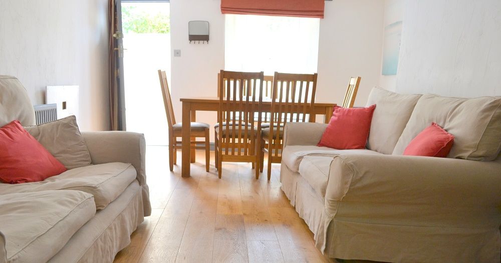 A cozy sitting room with two beige sofas, red cushions, a wooden dining table and chairs, and a wooden floor.