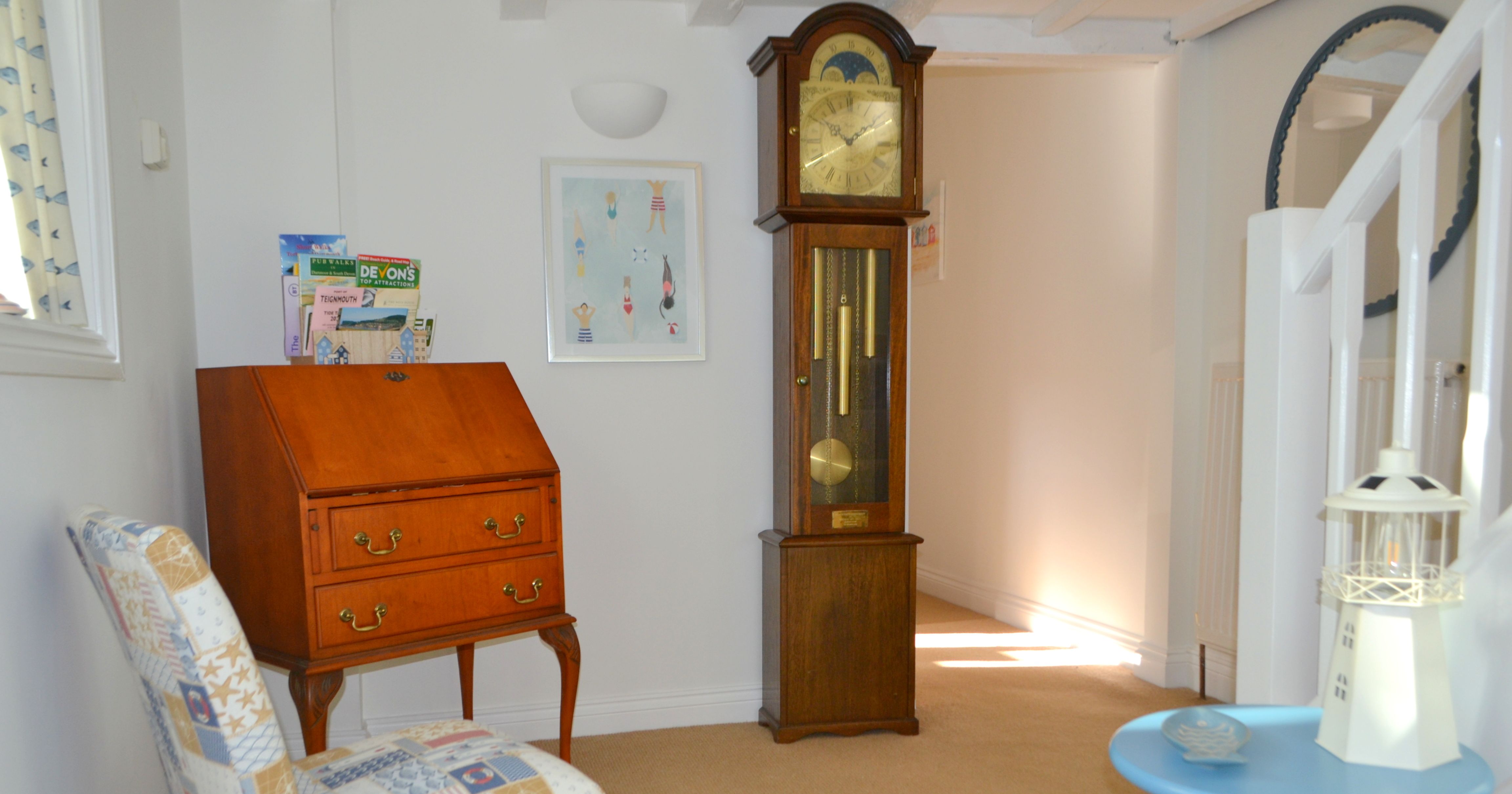 A cozy corner of a home interior featuring a grandfather clock, a wooden writing desk with magazines on top, a patterned upholstered chair, and a blue side table with a decorative lighthouse.
