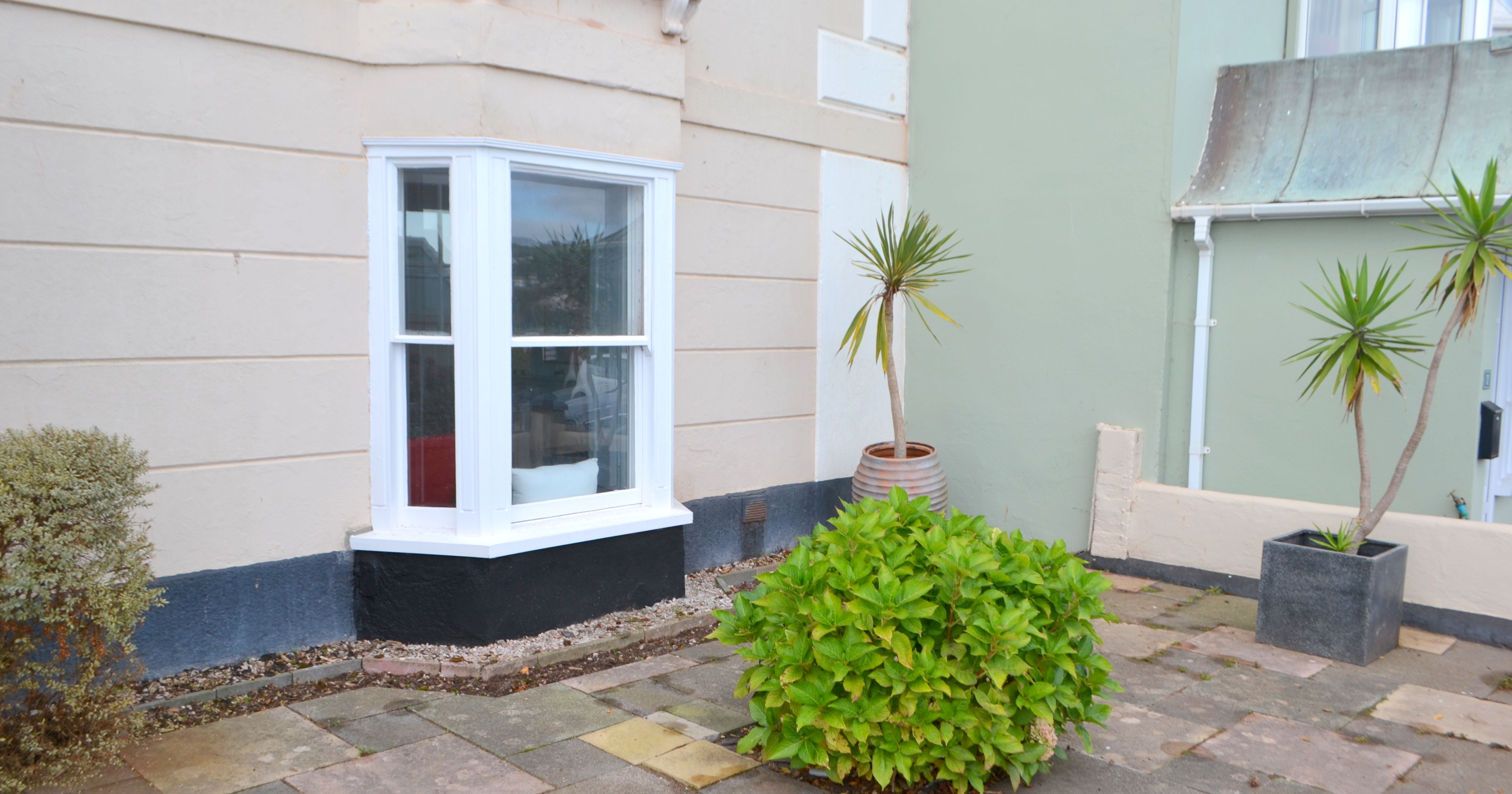 Small bay window on the ground floor of a house with a paved patio, potted palm plants, and a green bush.
