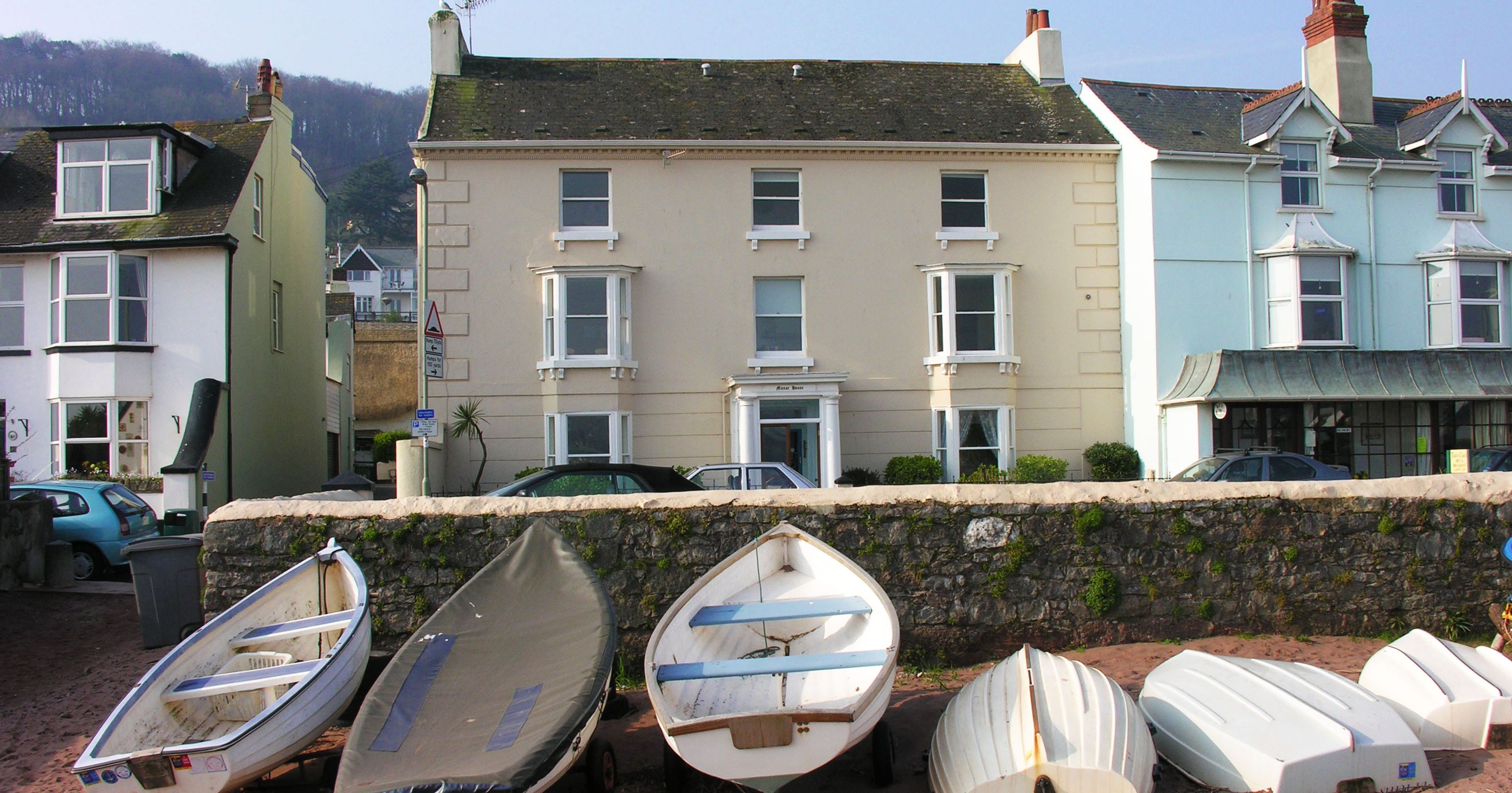 Row of small boats on sandy beach with large manor house and other buildings in the background