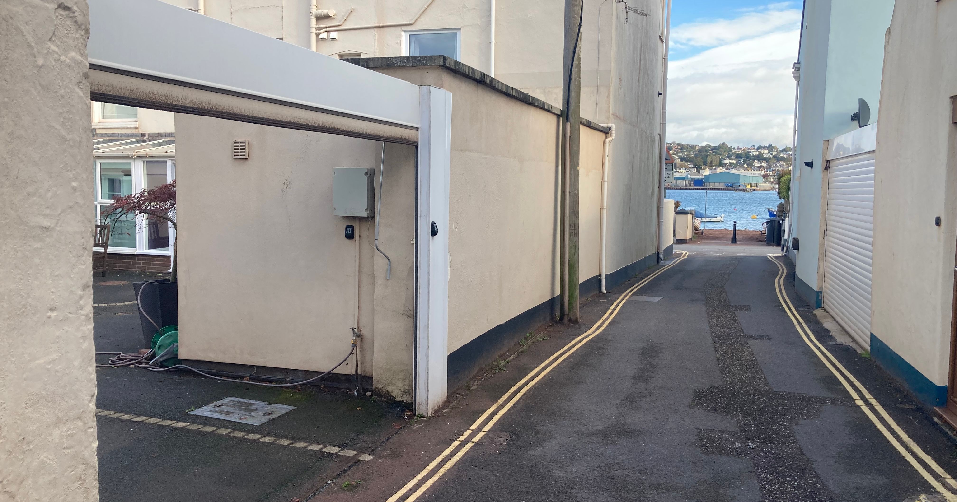 A narrow alleyway between buildings leading to a waterfront with distant houses and blue sky.
