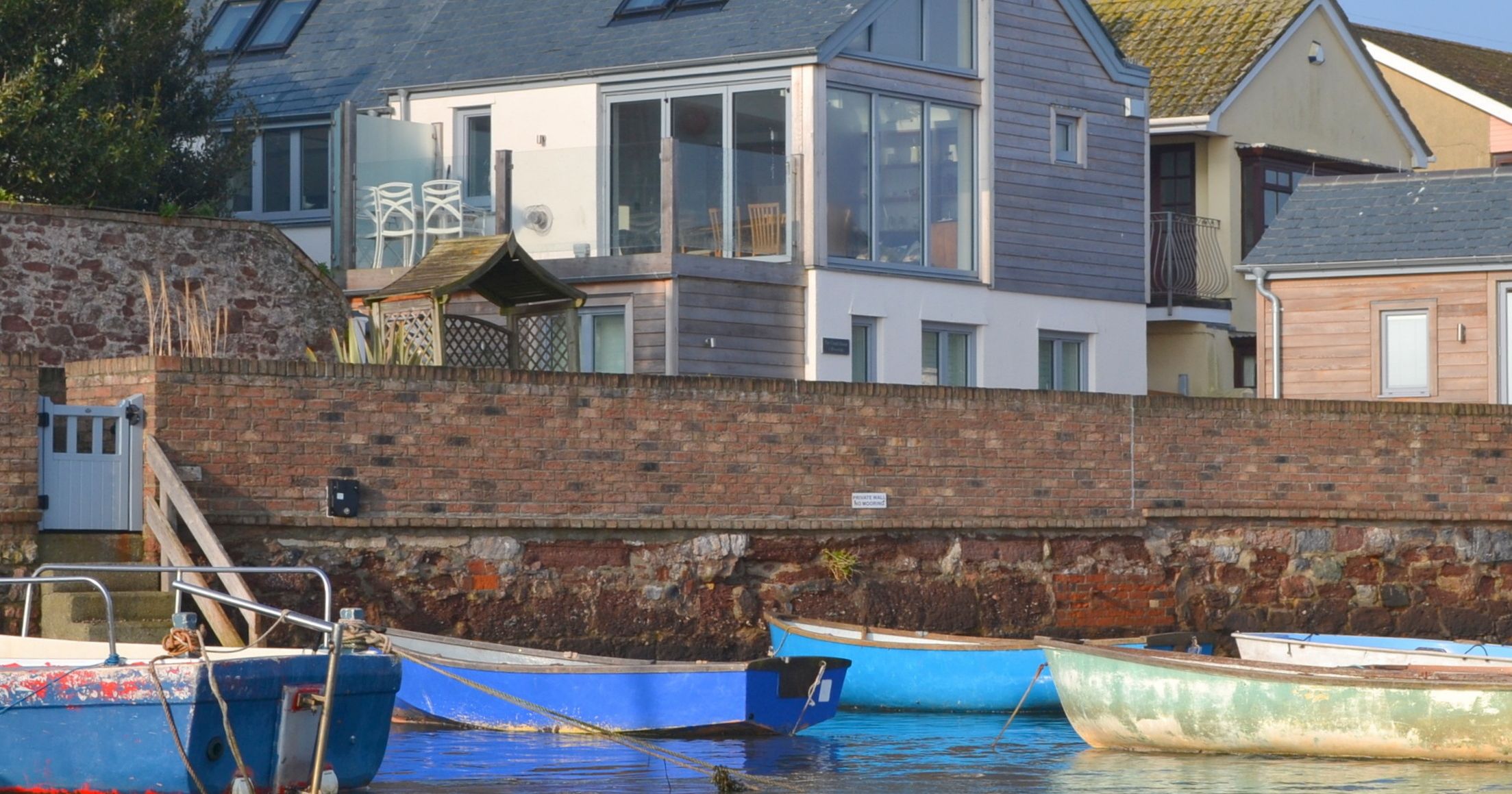 Modern house with large windows overlooking a calm waterfront with several small boats moored.