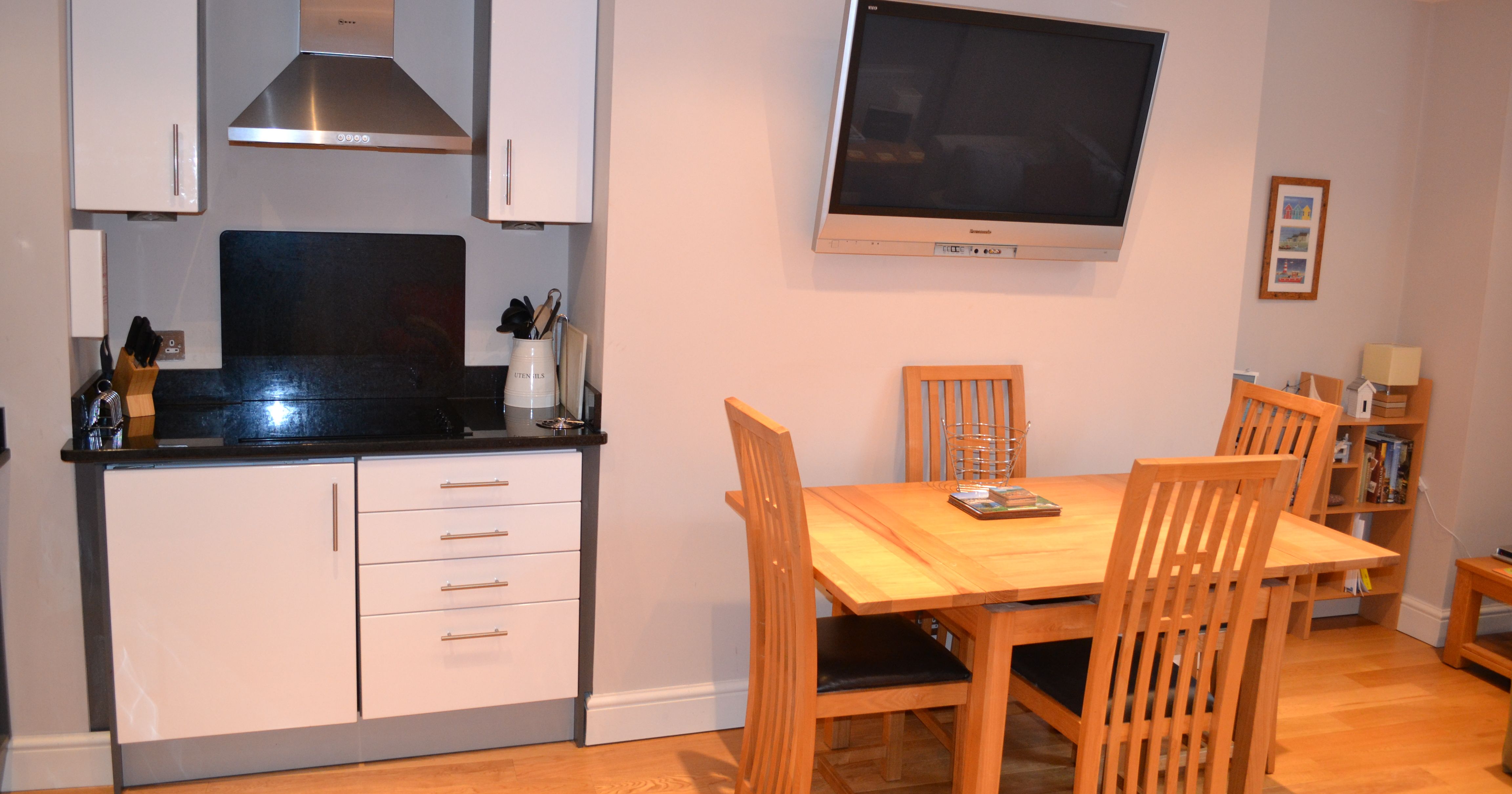 Modern kitchen and dining area with a wooden table, four chairs, flat-screen TV on the wall, and small kitchenette.