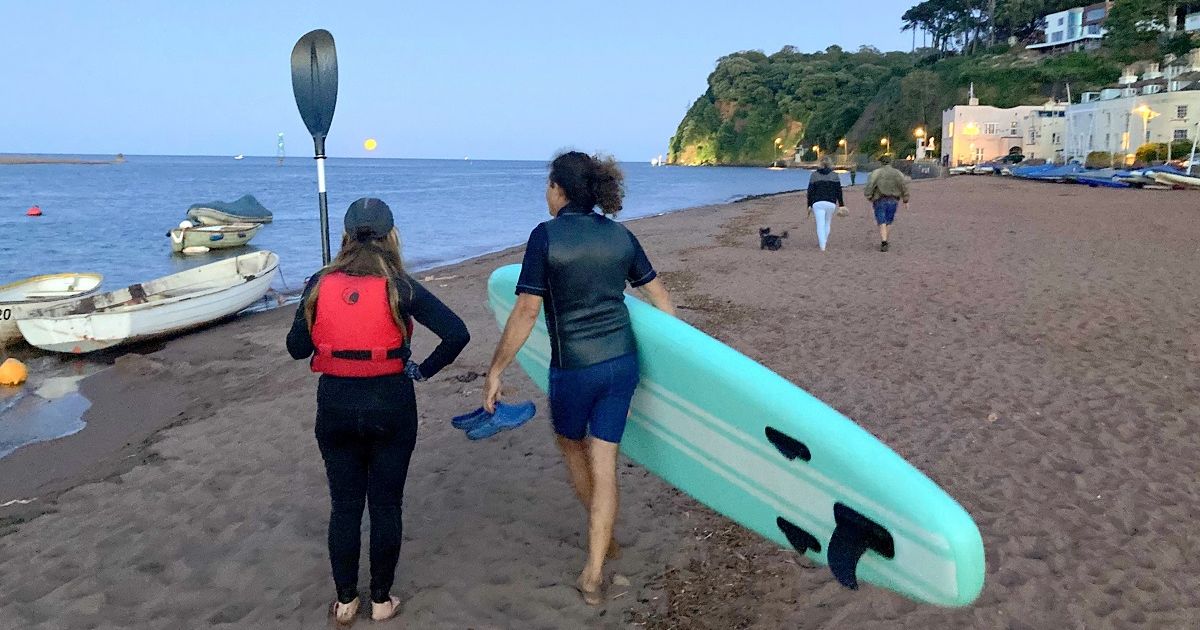 People walking along Shaldon Beach with paddleboards and boats at sunset with a full moon rising over the water.