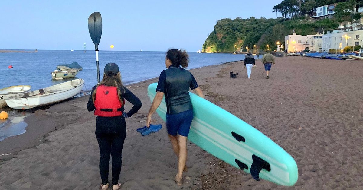 People walking along Shaldon Beach with paddleboards and boats at sunset with a full moon rising over the water.