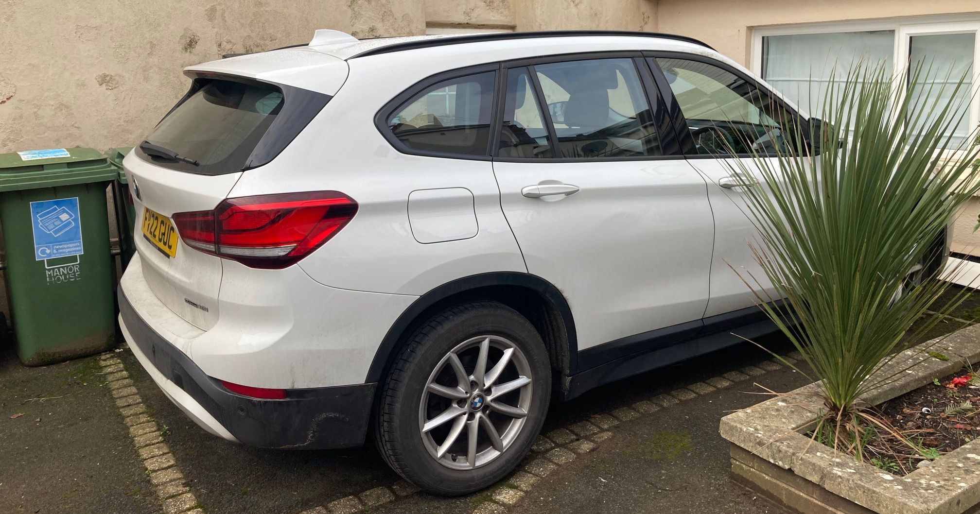 A white SUV parked in a small parking bay next to a green wheelie bin and a low building.