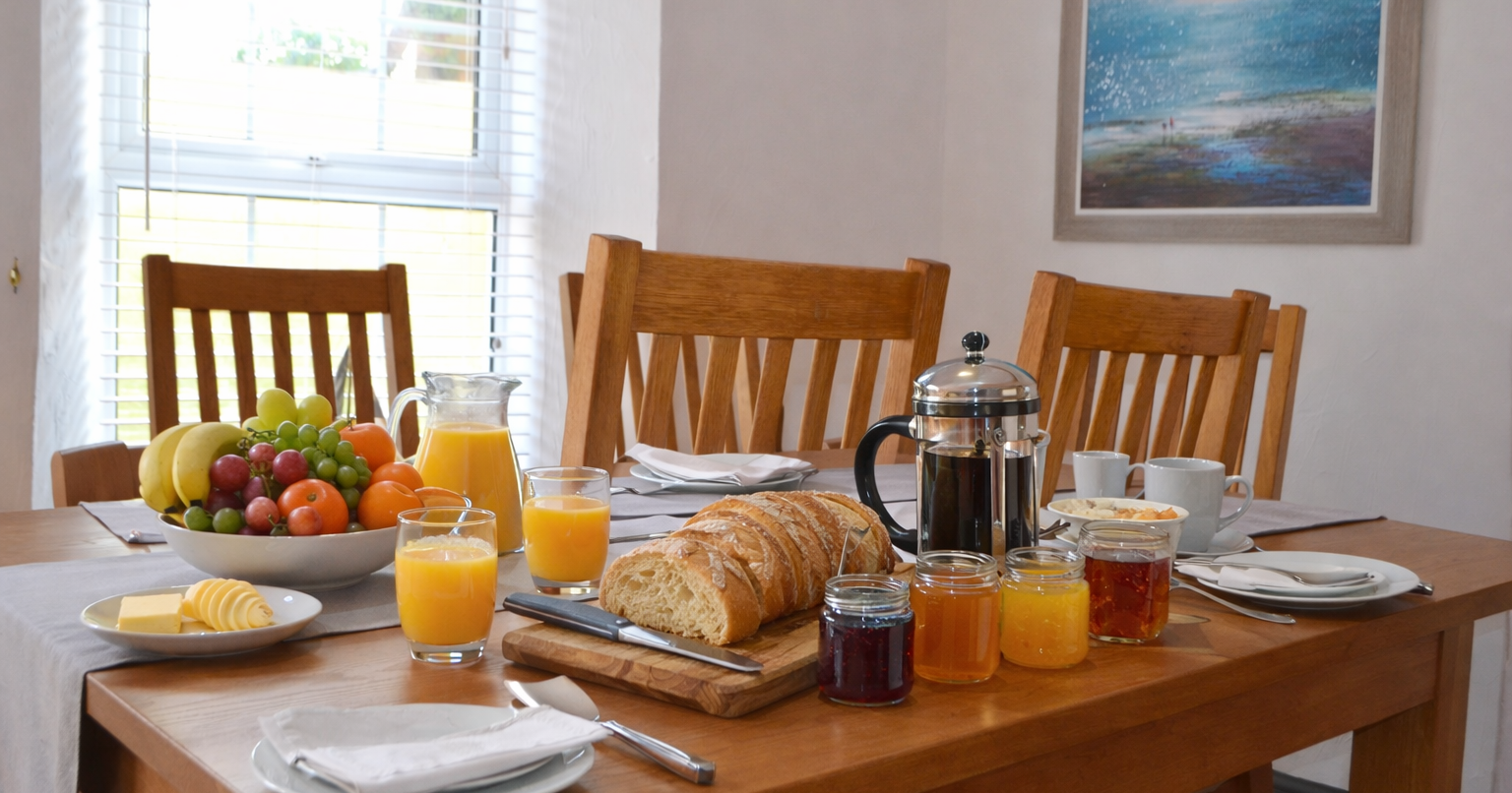 Continental breakfast with bread, fruit, juice, coffee, and jams on a wooden table in a bright room.
