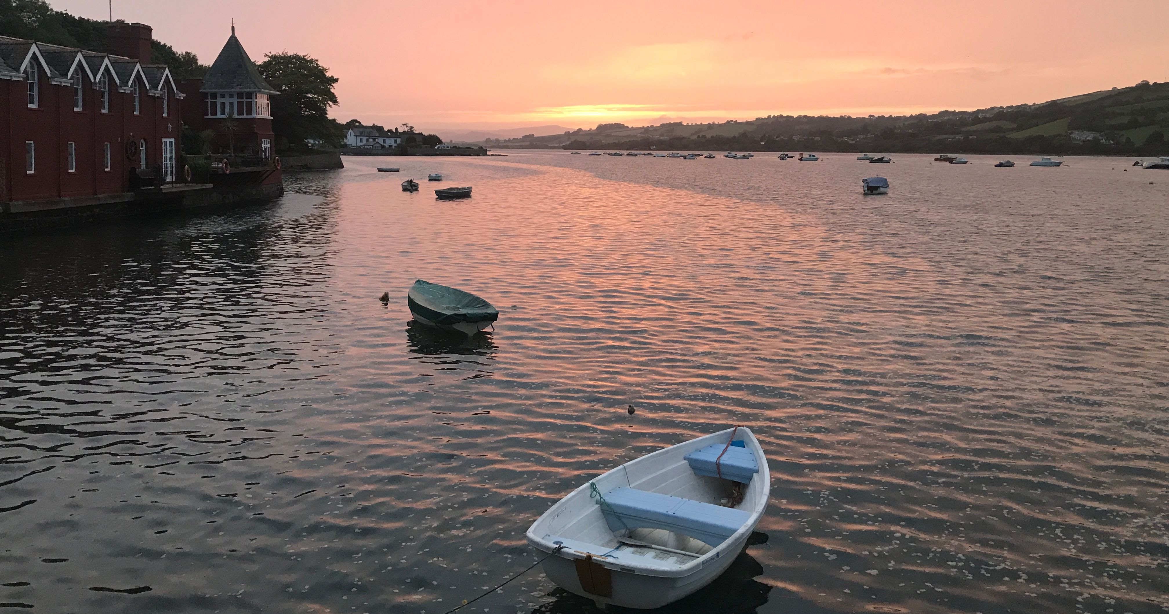A small rowboat floating on the River Teign at sunset with red riverside buildings and distant hills visible.