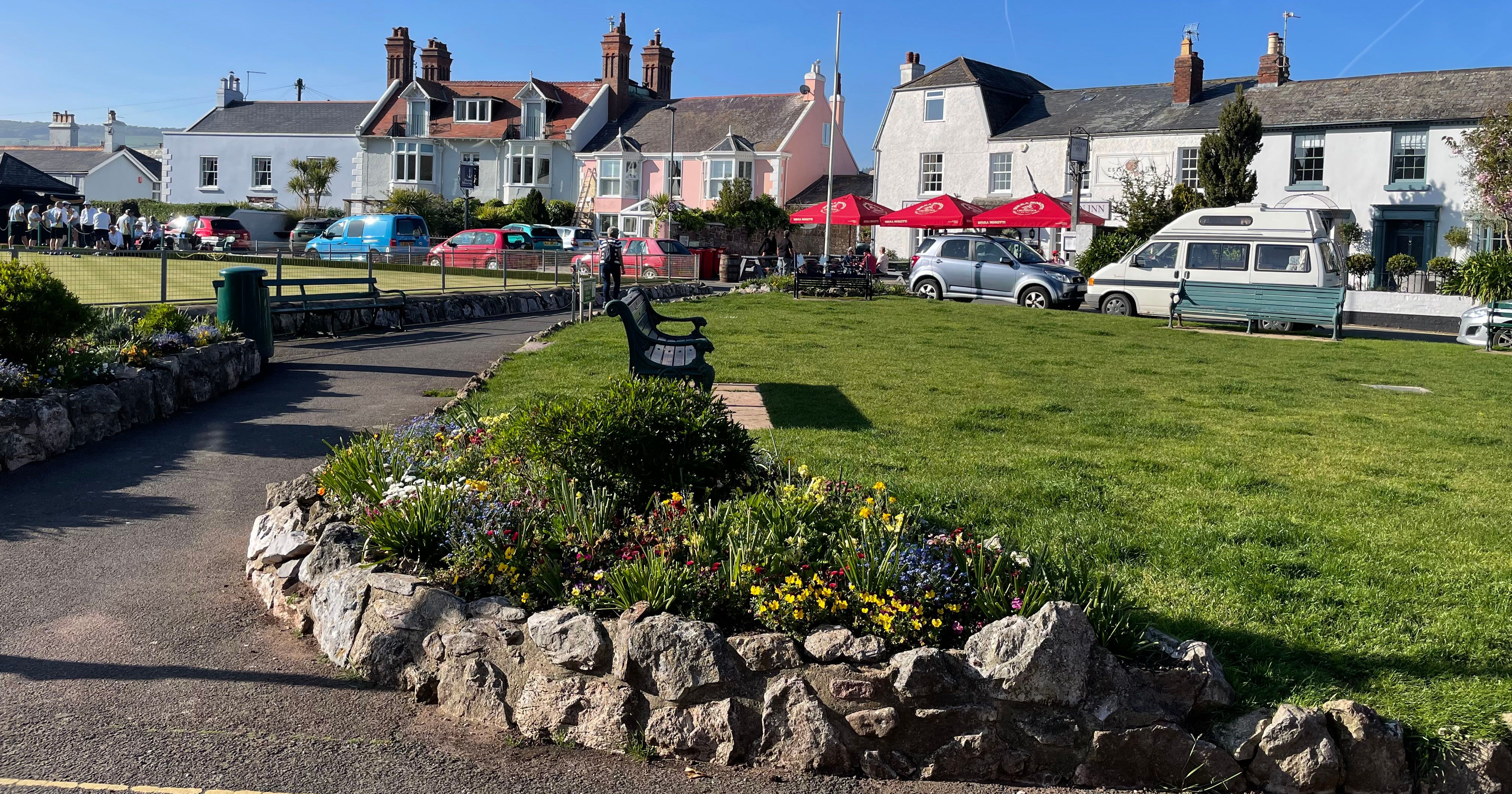 A sunny day at Shaldon bowling green, with people playing bowls, benches, flower beds, and colorful houses in the background.
