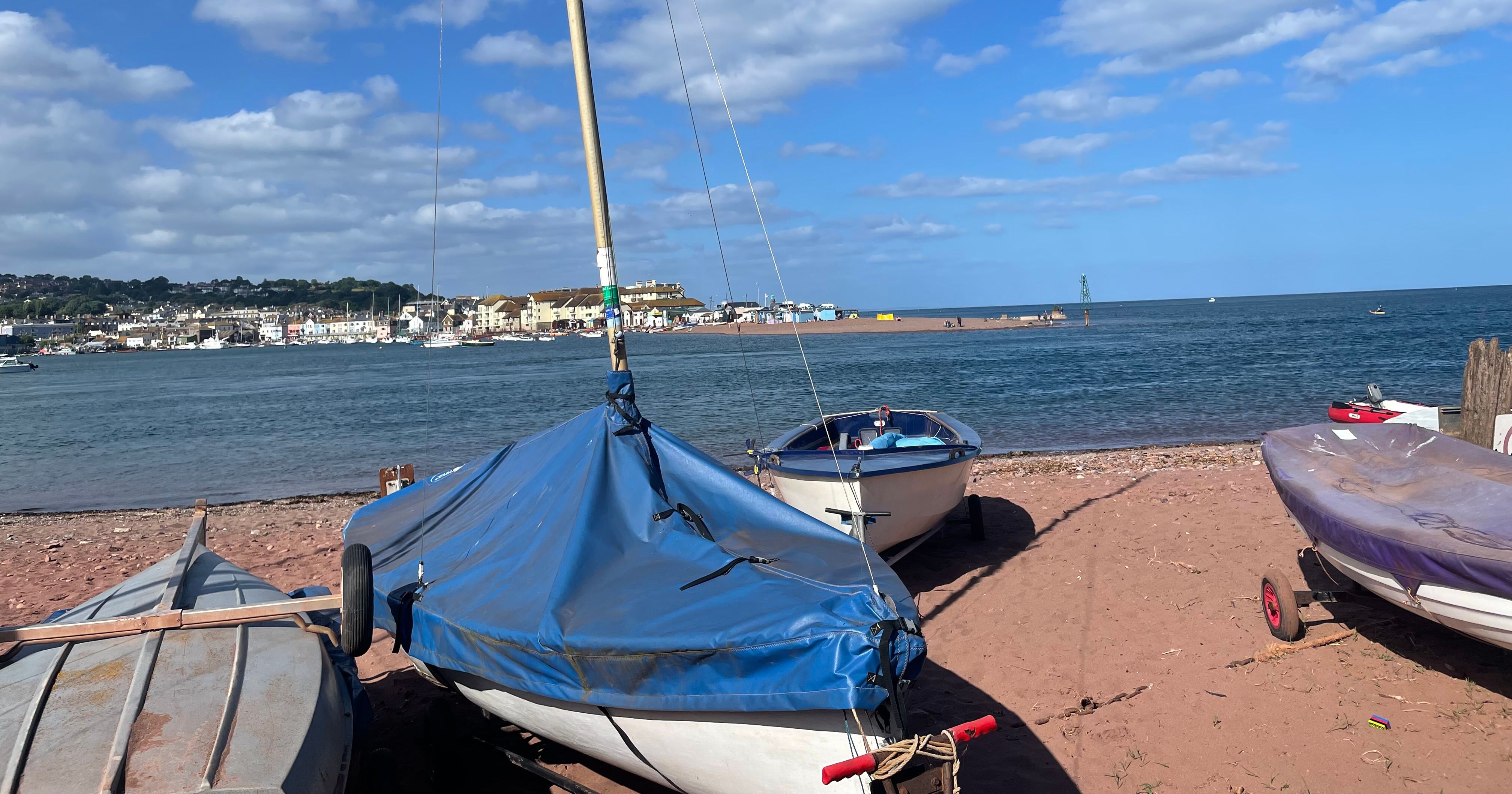 Small sailboats resting on a sandy beach with buildings and hills visible across a river estuary under a blue sky with scattered clouds.