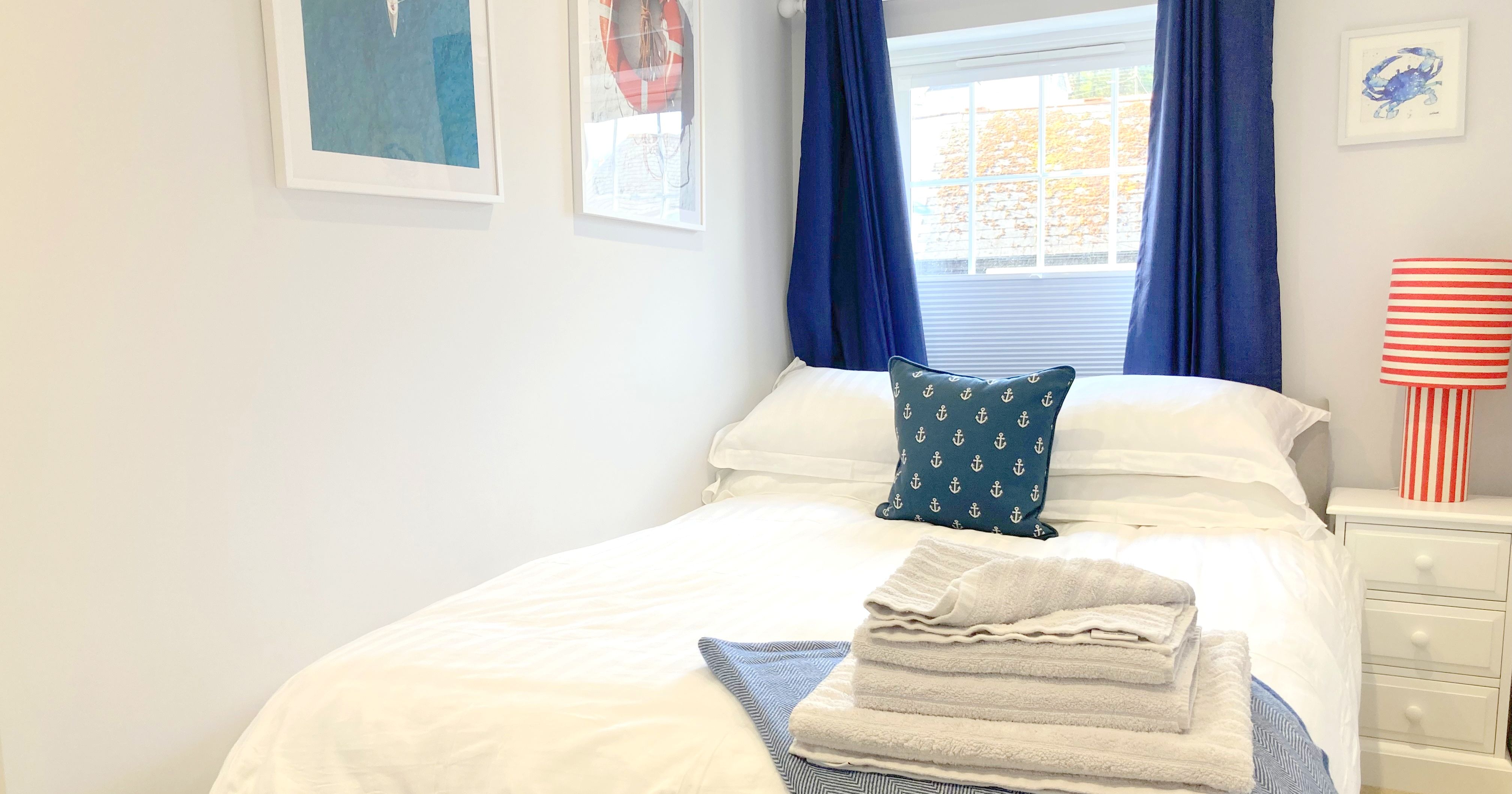 Cozy double bedroom with nautical decor, blue curtains, and neatly folded towels on the bed.