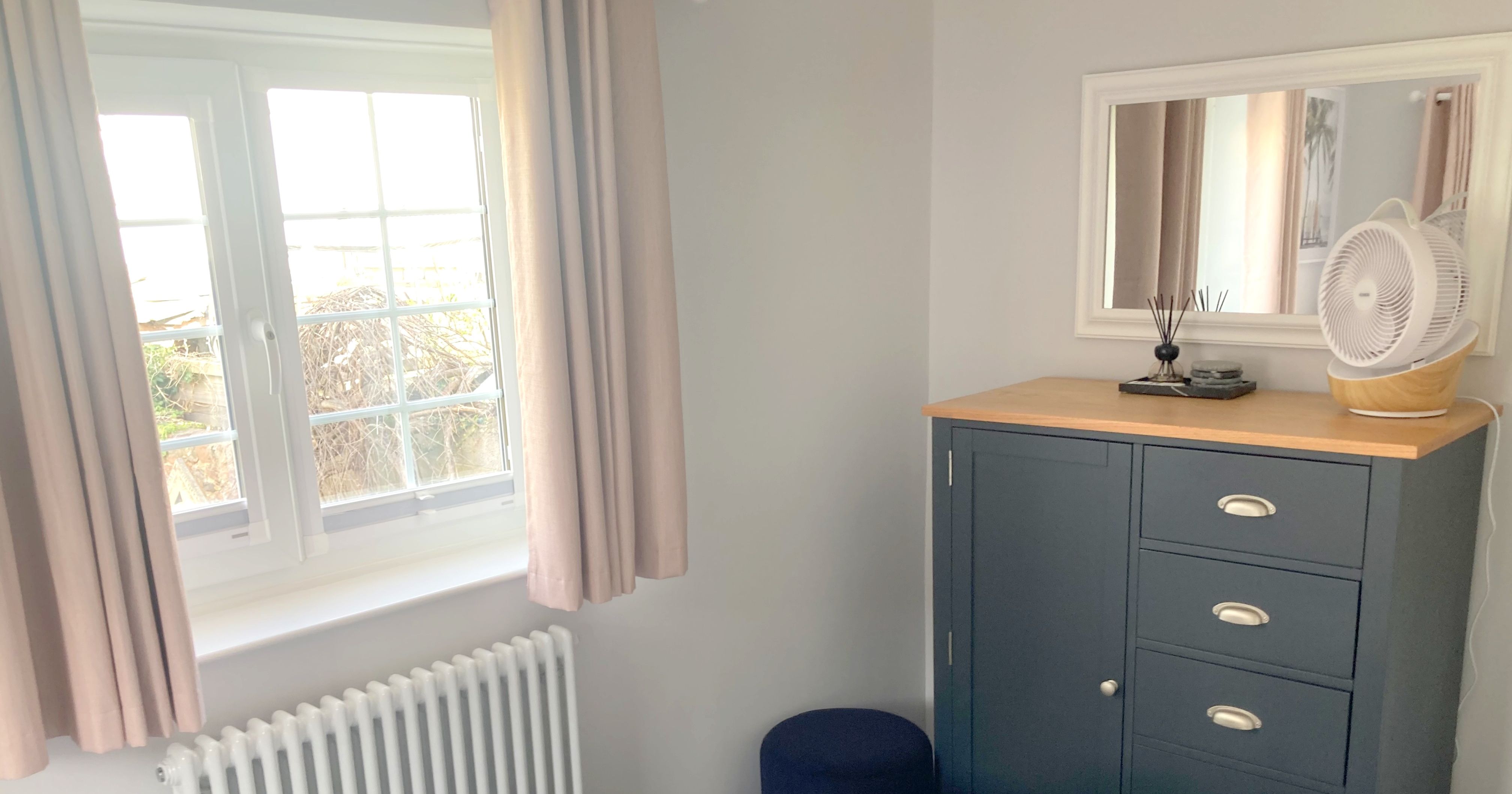 Bedroom corner with a dark blue chest of drawers, a fan, and folded towels on a bed near a window with curtains.