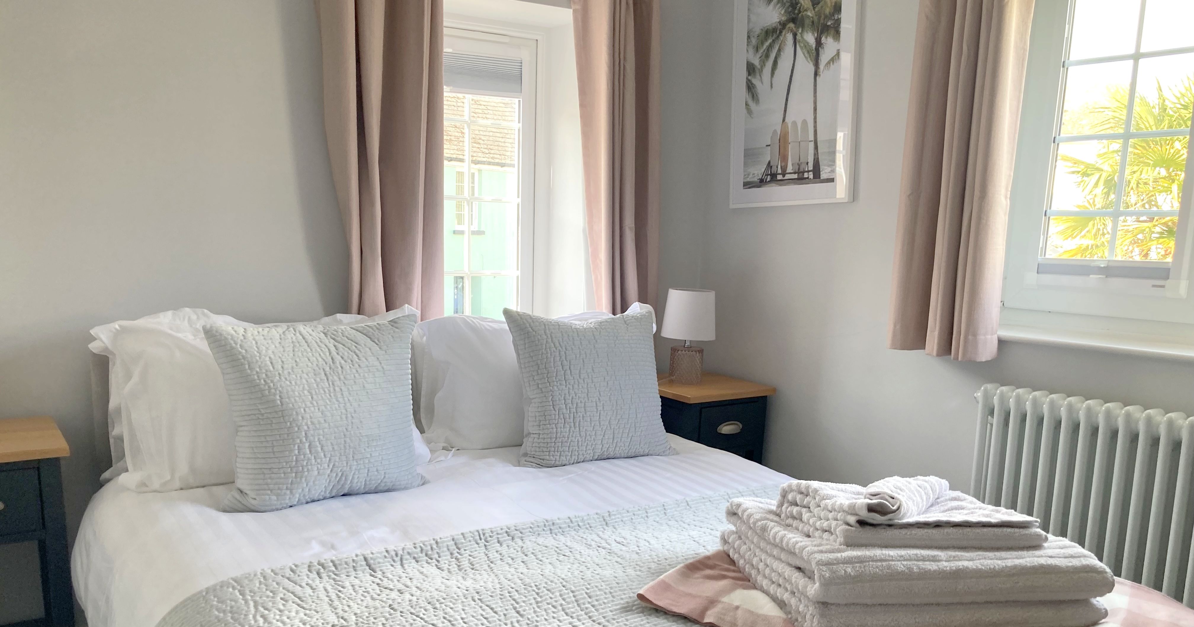 Bright, neatly made bedroom with king-size bed, light bedding, two large pillows, stacked towels, side tables, and two windows with beige curtains.