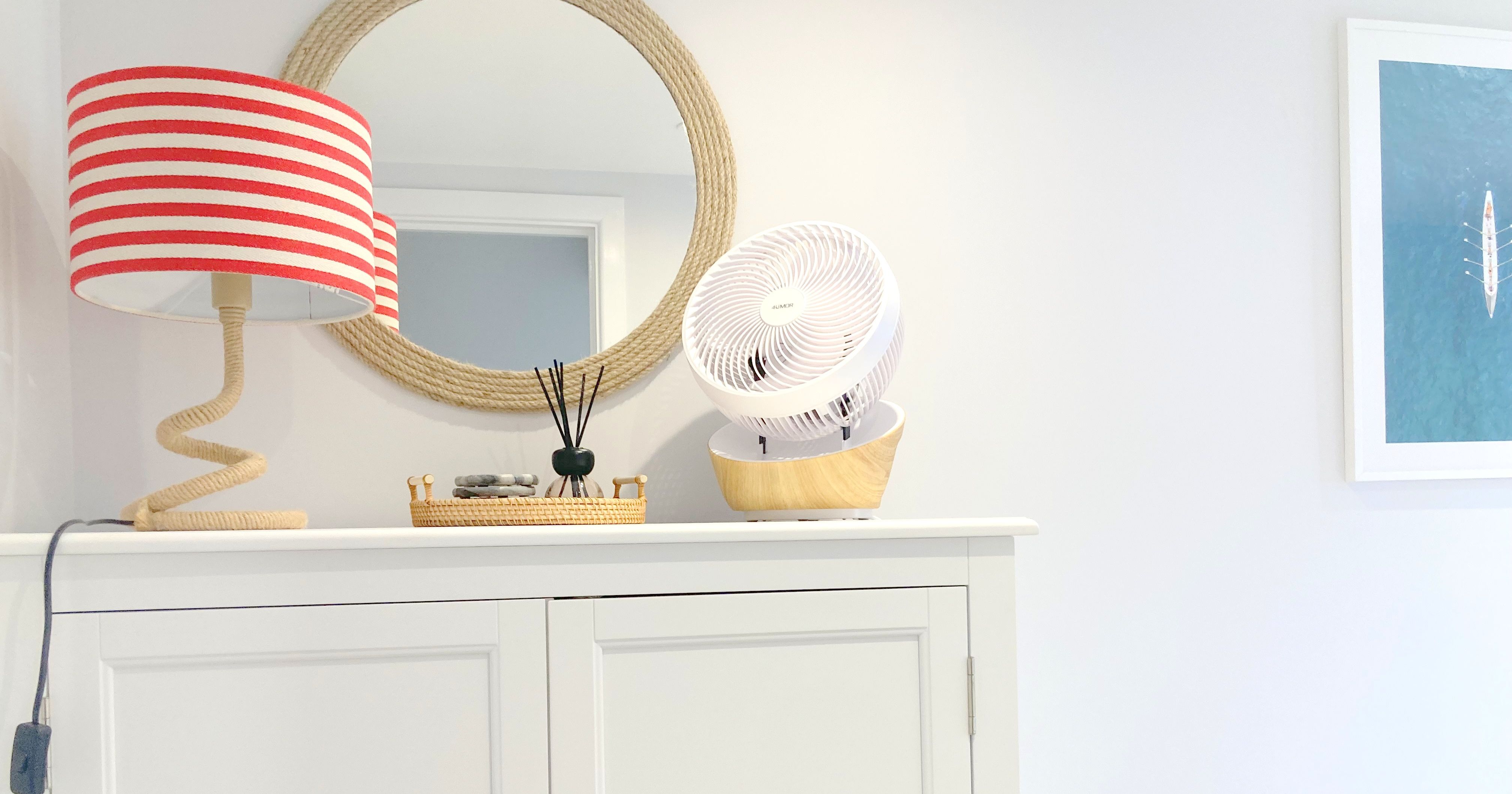 White storage cabinet with a red and white striped lamp, round rope mirror, small fan, and decorative tray in a bright, coastal-inspired room.