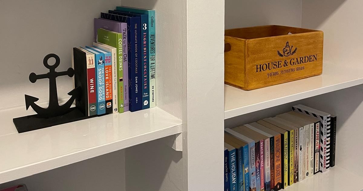 A white bookshelf with various books, a wooden box labeled 'House & Garden', a black anchor bookend, a starfish decor, a brown ceramic jug, and some puzzles.