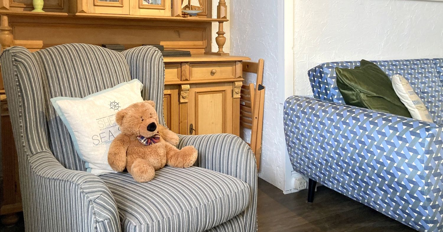 A cozy living room scene featuring a plush teddy bear on a striped armchair, with a wooden hutch, a blue patterned sofa, and decorative pillows.