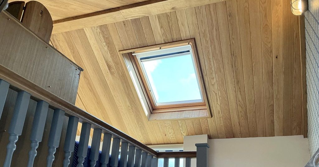 A wooden staircase leading up to a skylight window set in a slanted wooden ceiling.