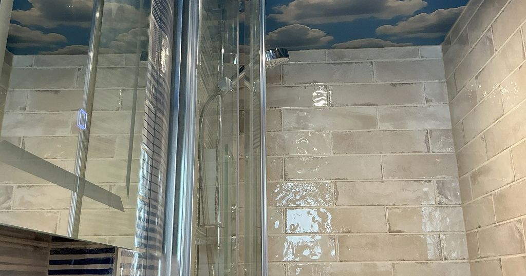 Shower stall with glass doors, beige tiles, and a ceiling painted with clouds on a blue sky.